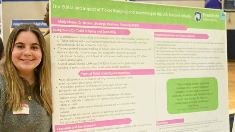 A woman stands at left next to a large research poster on a table.