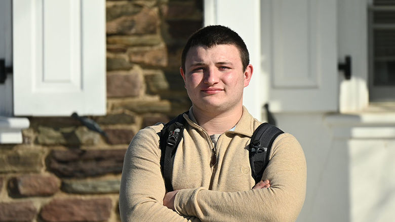 A man wearing a backpacks stands against a stone building with his arms crossed over his chest.