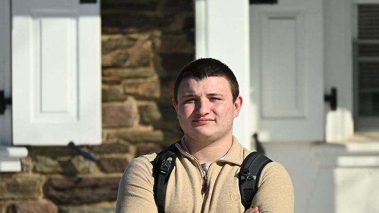 A man wearing a backpacks stands against a stone building with his arms crossed over his chest.