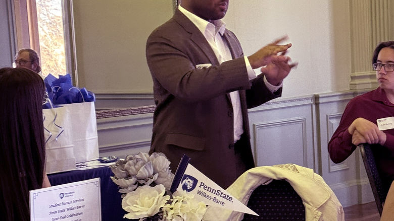 A man standing aside a table gestures while speaking.