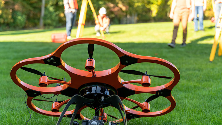 A drone on the grass with surveying equipment and people in the background.