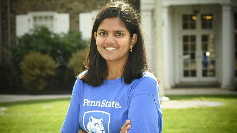 A student wearing a blue shirt with the Penn State mark, standing in front of a historic building.