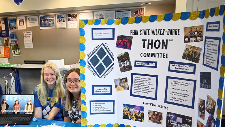 Two women at a table sitting next to a posterboard promoting the campus' THON organization.
