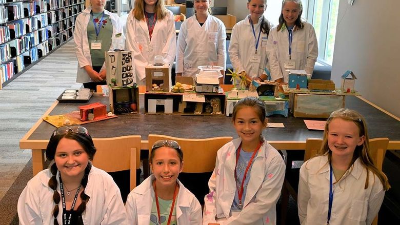 A group of young students in lab coats posing in two rows in front of and behind a table.