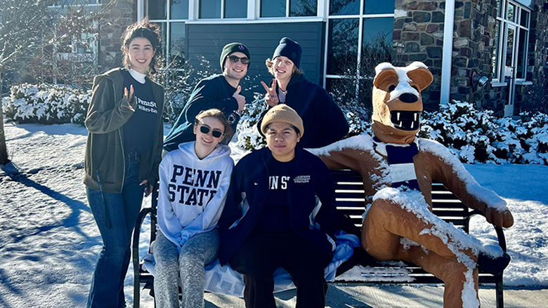 Students sitting on a bench with the Nittany Lion mascot statue, with snow in the background