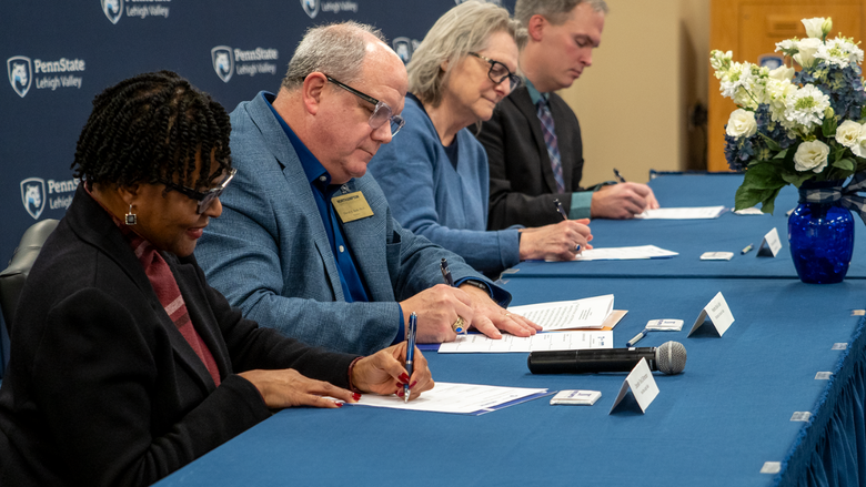 four individuals sit at a long table with their heads down and sign a document 