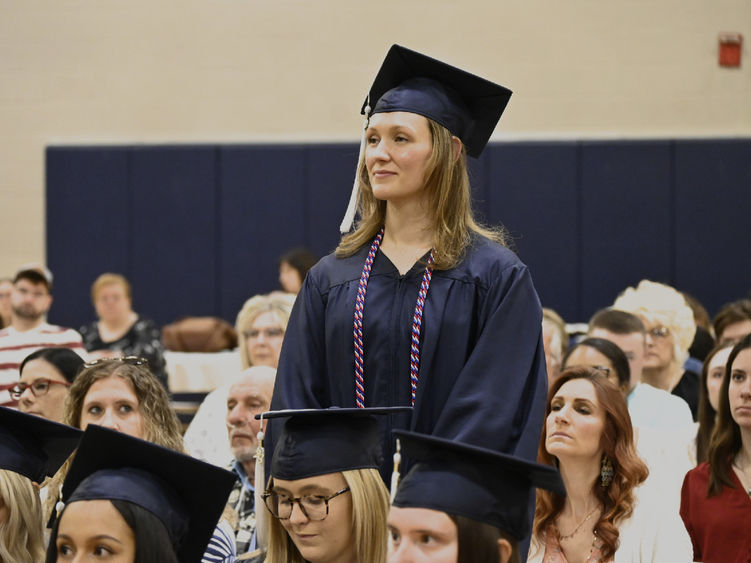 A woman wearing a cap and gown as well as a cord indicating her veteran status stands among seated graduates and audience members