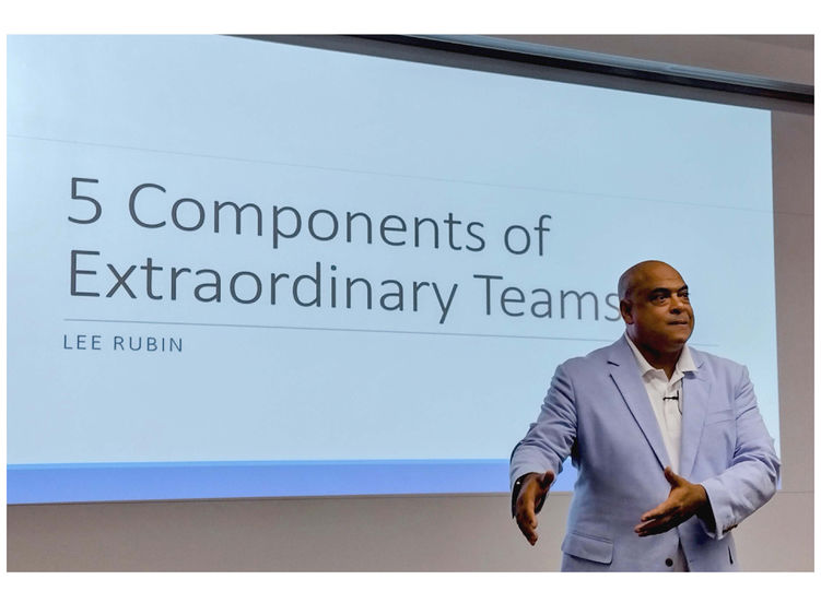 Man in front of screen speaking to audience, the screen displays "5 Components of Extraordinary Teams"