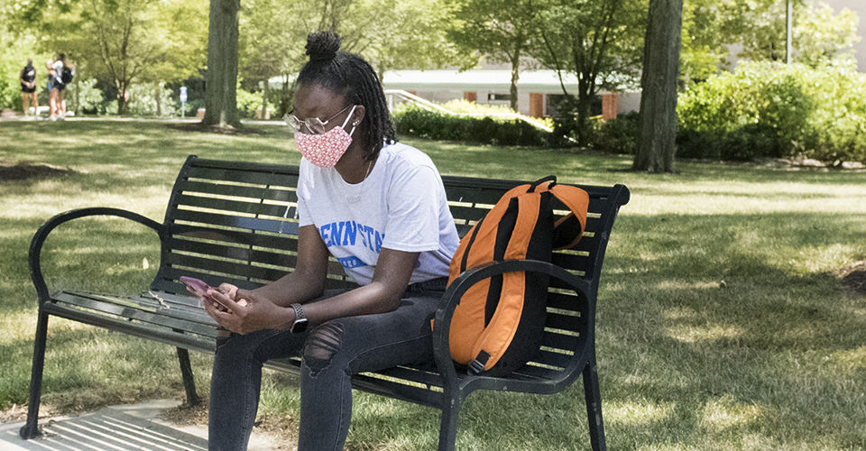 A Penn State student wearing a face mask looks at her phone while sitting on a campus bench.