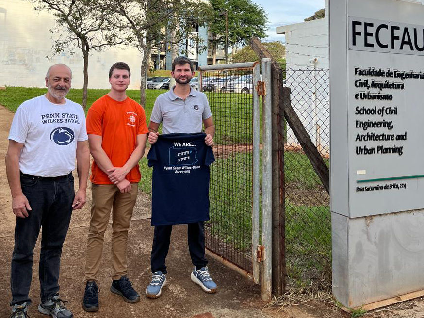 Three people, one holding a Penn State Wilkes-Barre shirt, stand outside a building in Brazil.