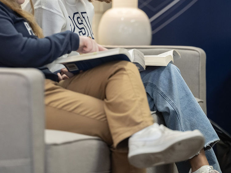 An image showing the legs of two students with textbooks on their laps, as one student gestures at a book.