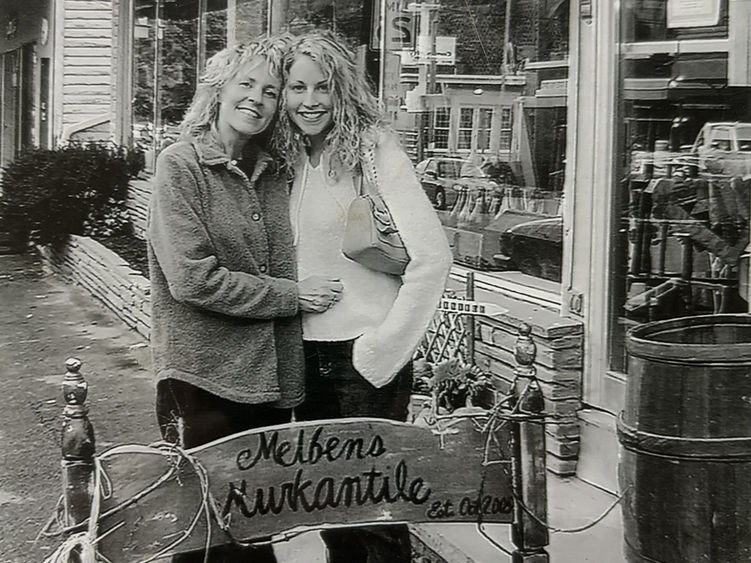 A black and white photo of two woman standing outside a business with their arms around each other.