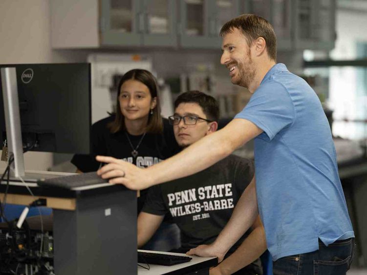 A man standing at right assists two seated students working on a computer.