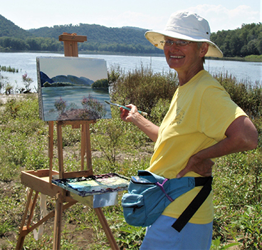 Artist Sue Hand with a paintbrush in her hand standing in front of a canvas on an easel. In the background is the Susquehanna River.